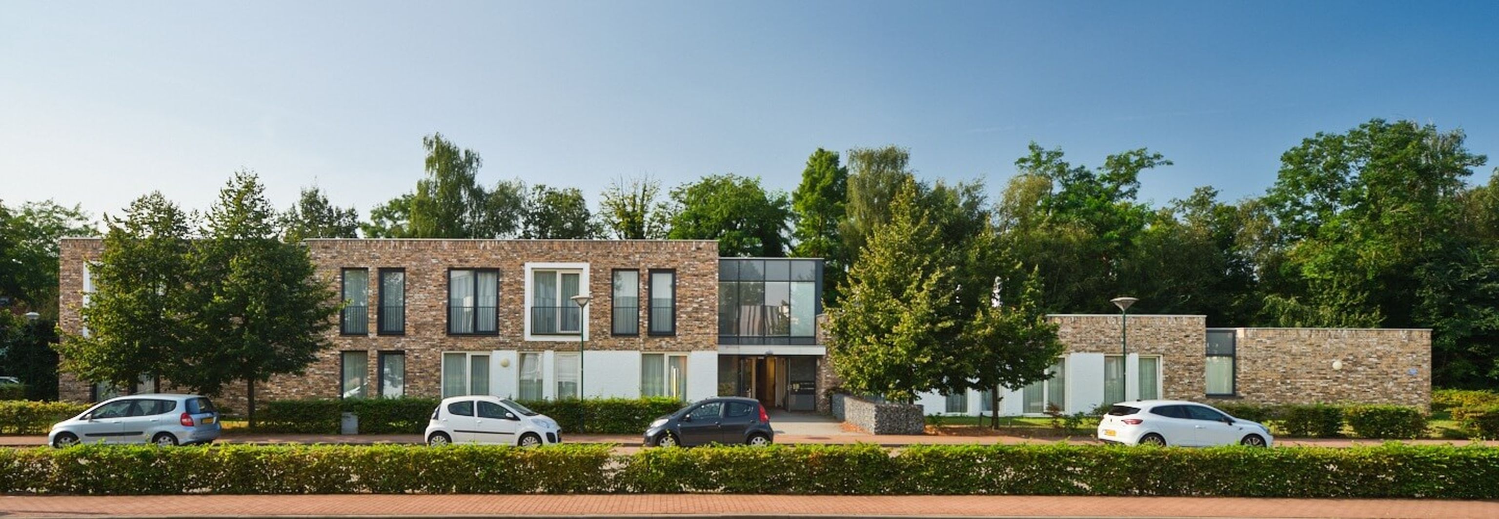 Low two-story brick building with large windows and glass entrance, trees and hedges, and cars parked in front.