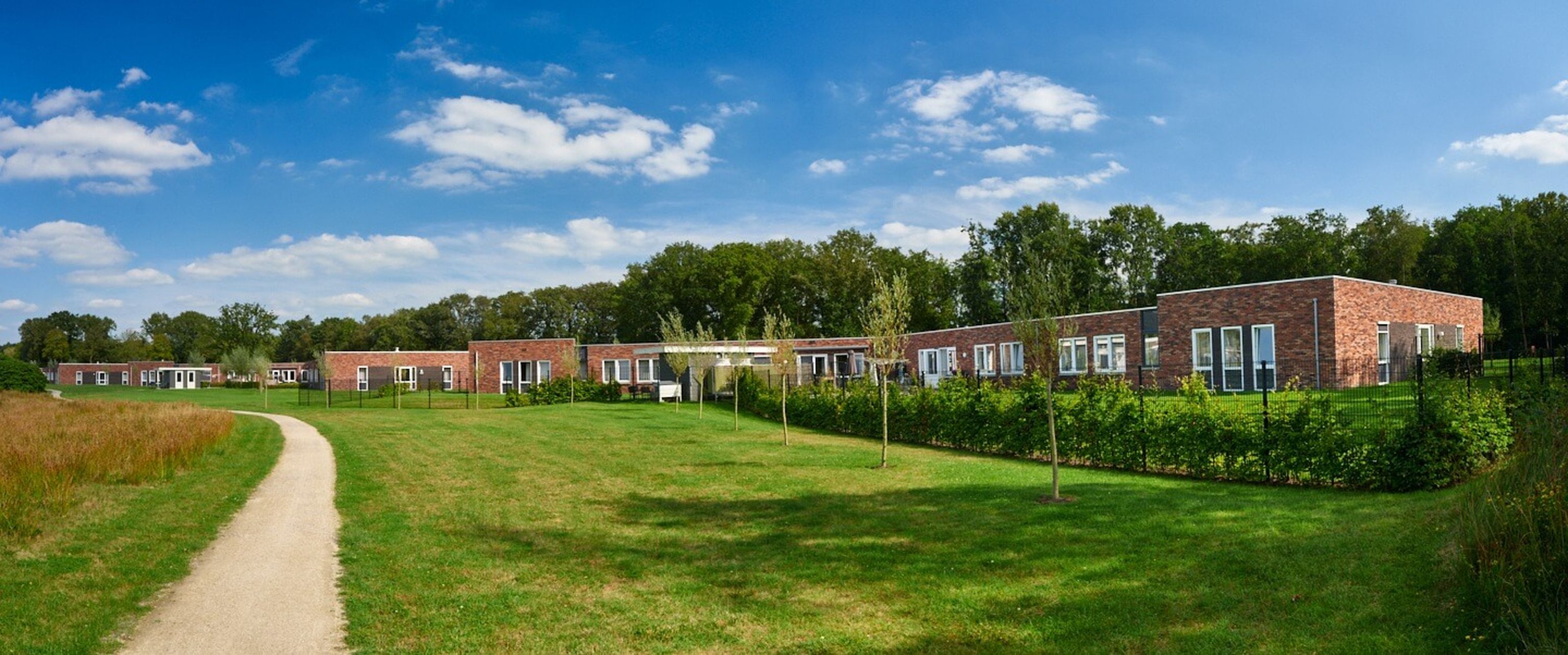 Panoramic view of single-story brick homes along a walking path, with manicured lawns, young trees and a blue sky with scattered clouds.