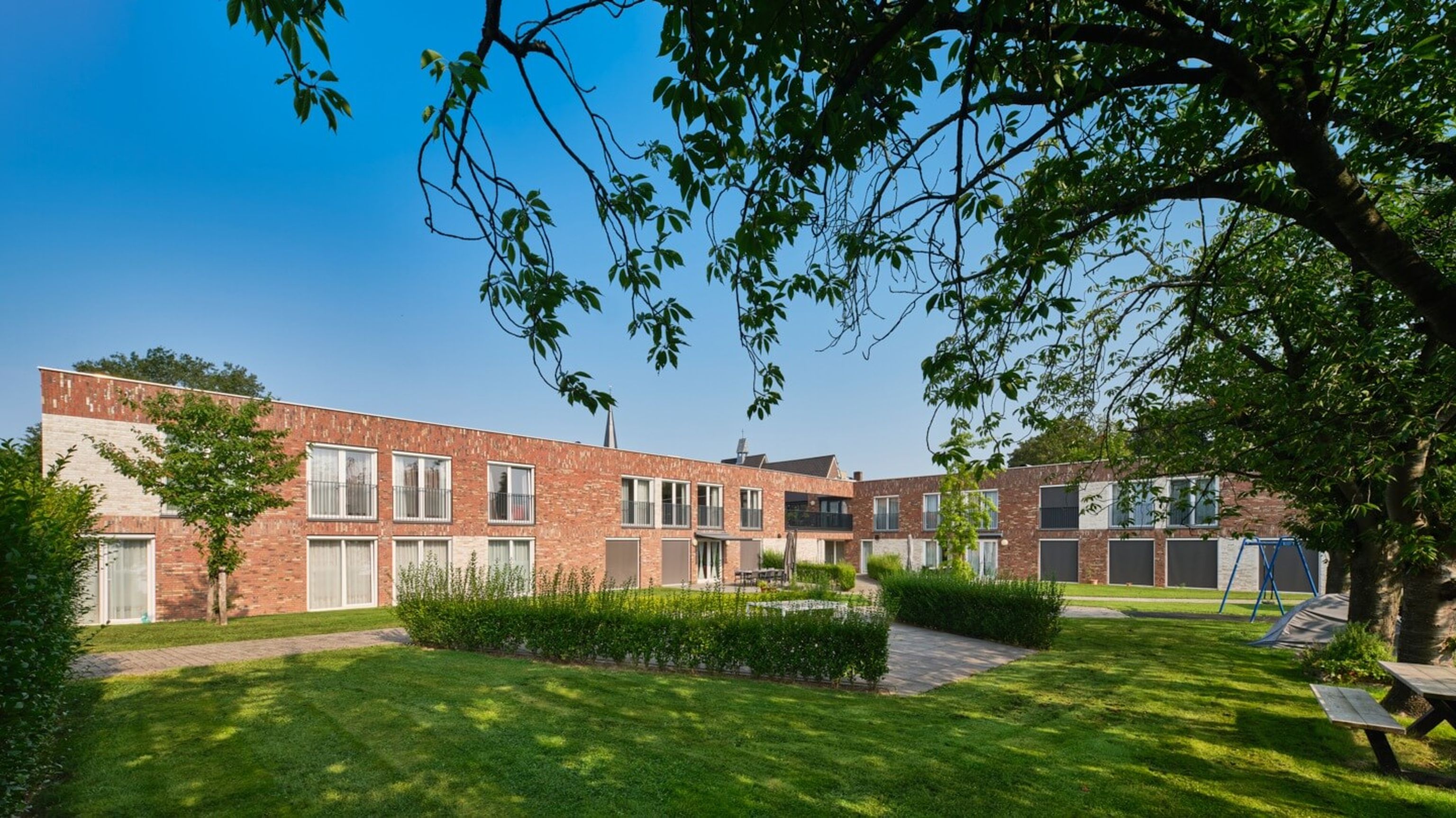Two-story red-brick apartments around a green courtyard with hedges, trees, swing set and picnic bench beneath a clear blue sky.