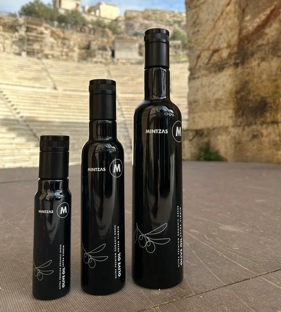Three Miron Glass Mintzas olive oil bottles of varying heights on a textured surface, with ancient stone steps in the background.