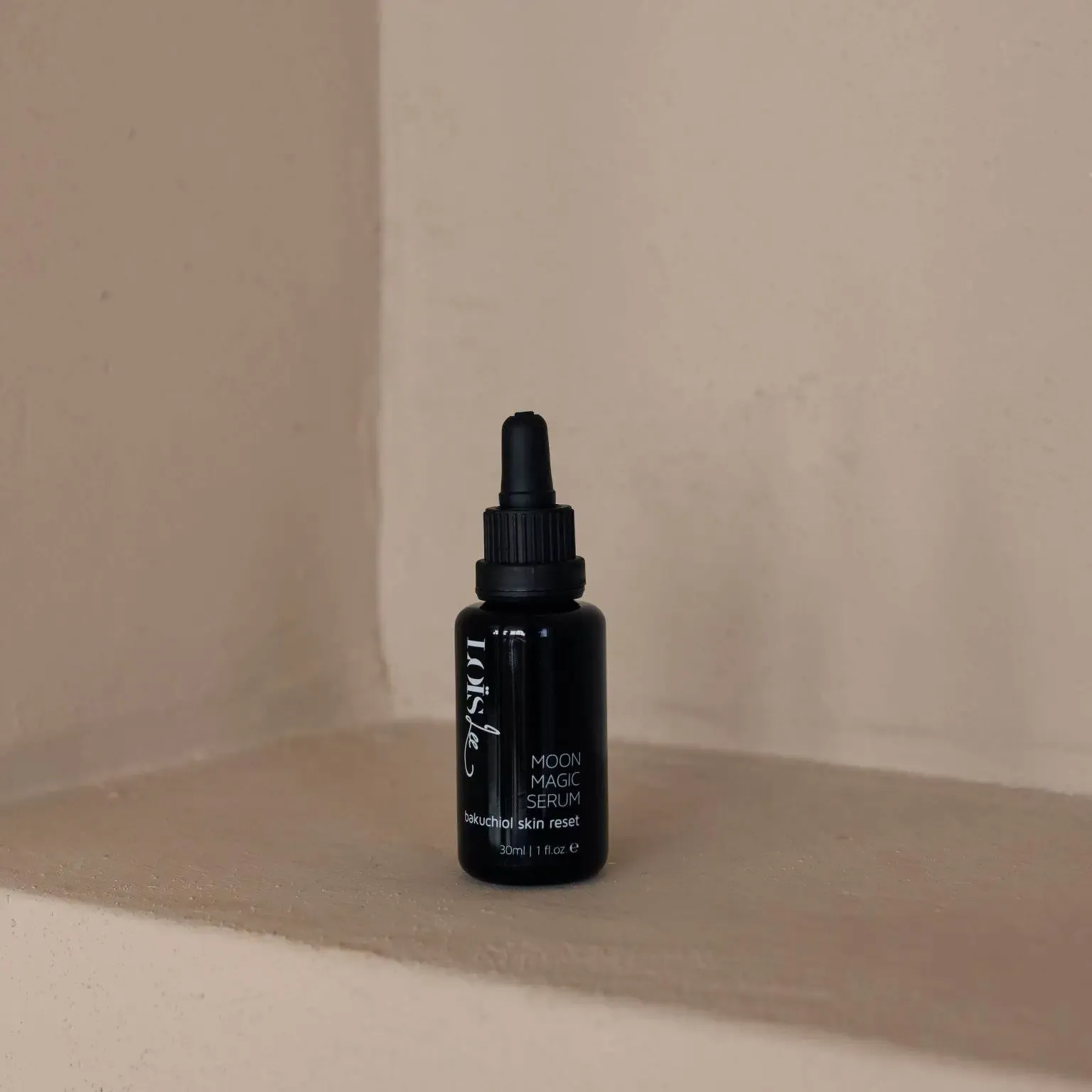 A small dark Miron dropper bottle labeled Moon Magic Serum sits on a beige surface with a pale beige wall in the background.