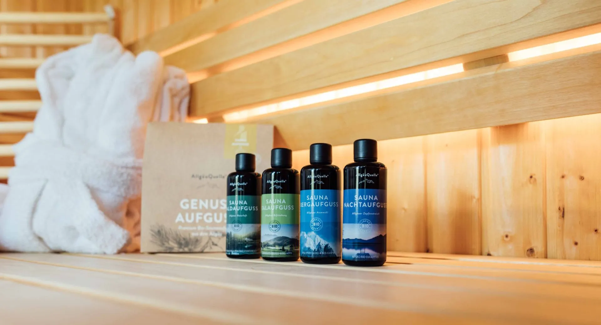 Five miron sauna bottles lined up on a wooden bench, beside a folded white towel and a cardboard box in a warm, sunlit sauna.
