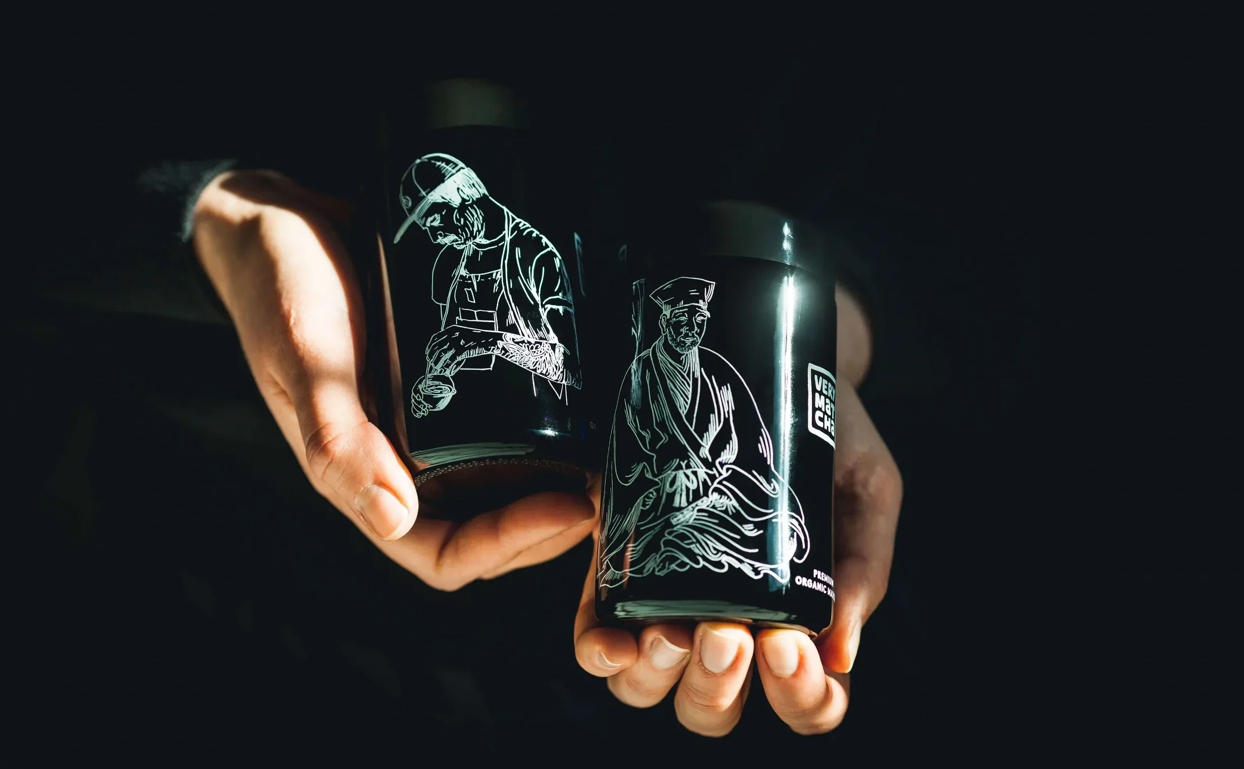 Two Miron Glass featuring white line-drawn illustrations of a craftsman and a seated sage, held by hands against a dark background.