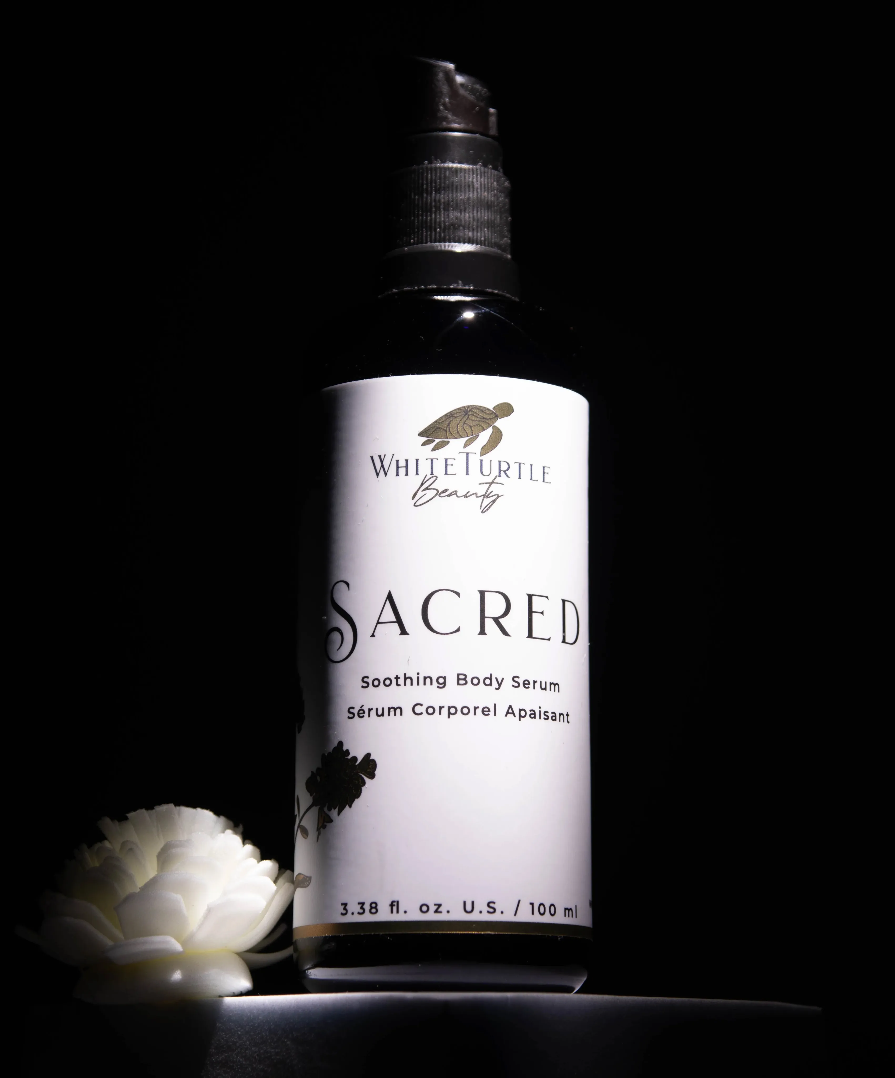 Close-up of a violet pump bottle labeled White Turtle Beauty Sacred Soothing Body Serum, set against a dark background with a white flower nearby.