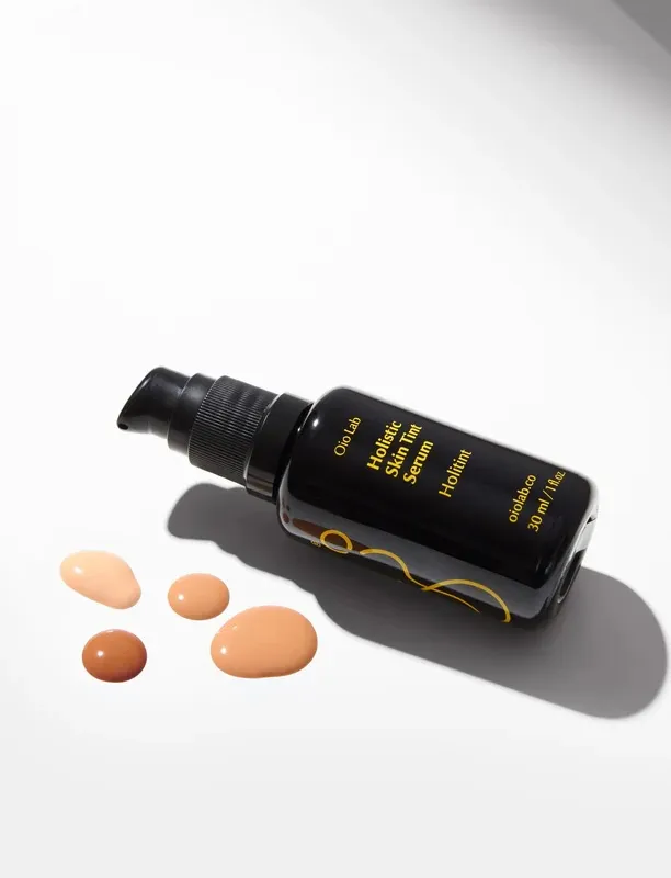 Miron glass bottle of Oil Lab Holistic Skin Tint Serum on a white surface, with several peach-colored serum droplets nearby.