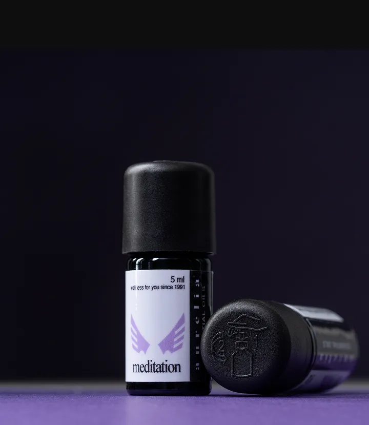 Small dark miron glass essential oil bottle labeled 'meditation' with a purple emblem, standing on a purple surface; another bottle lies on its side beside it.