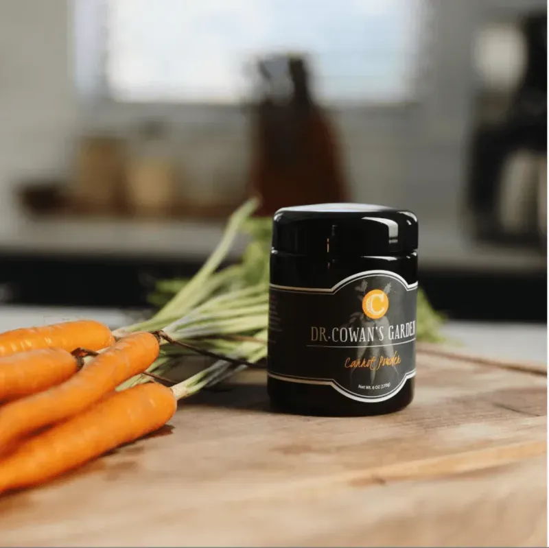 Miron jar labeled Dr. Cowan's Garden sits on a wooden surface next to fresh orange carrots; a blurred kitchen scene in the background.