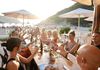 A group of people toasting at a seaside restaurant, sitting under umbrellas with a beach and sunset in the background.