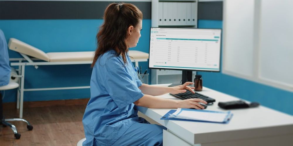 Nurse in blue scrubs working at a desk with a computer in a medical office; clipboard and small bottle nearby.