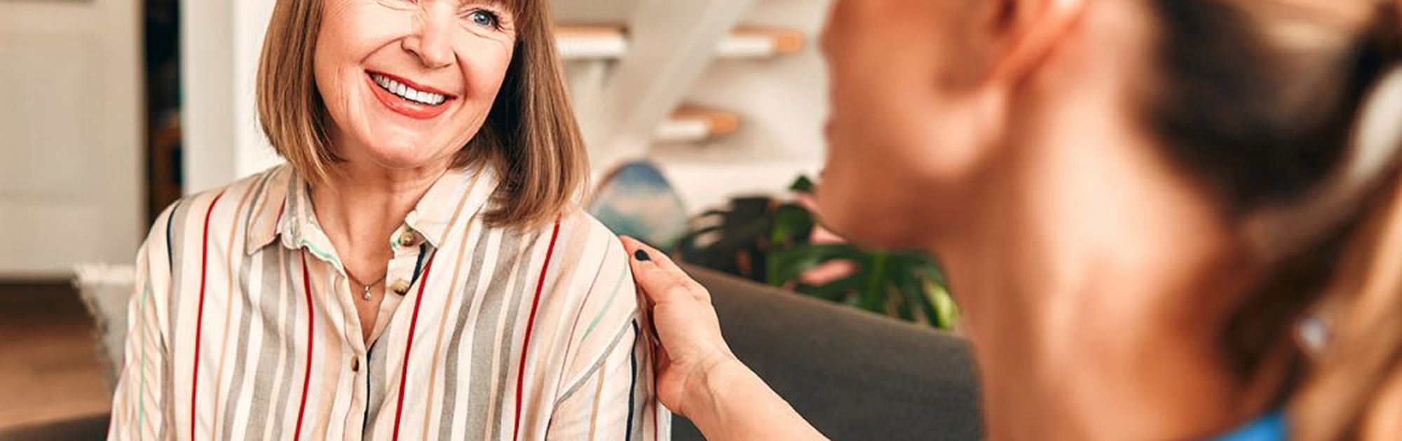 A woman with shoulder-length brown hair smiles while talking to another woman who is touching her shoulder, indoors by a staircase.