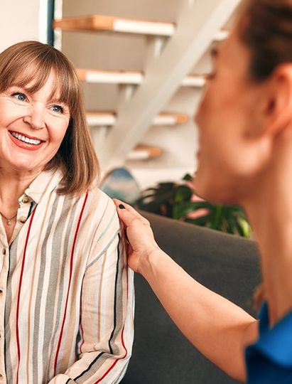 A woman with shoulder-length brown hair smiles while talking to another woman who is touching her shoulder, indoors by a staircase.