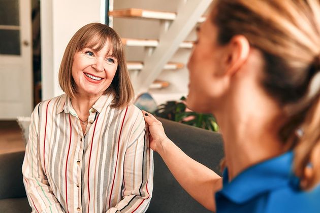 A woman with shoulder-length brown hair smiles while talking to a nurse who is touching her shoulder, indoors by a staircase.