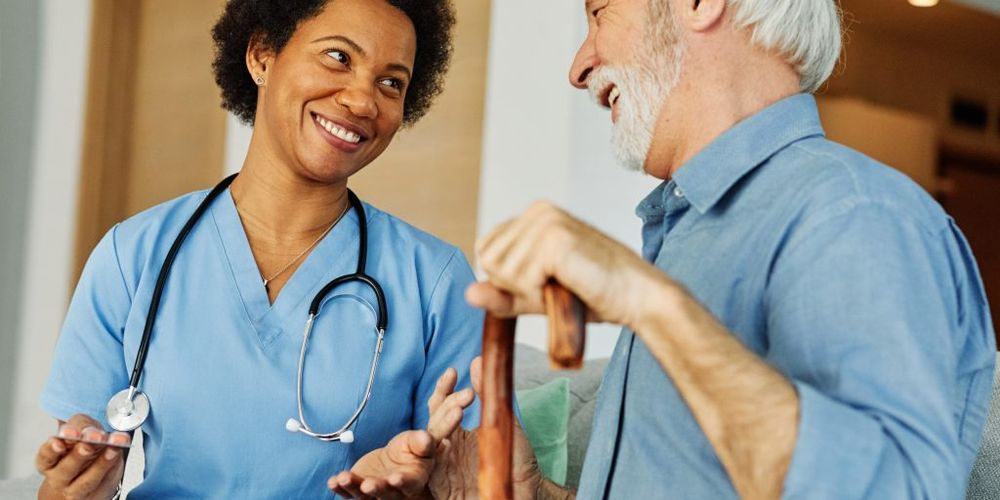 A nurse in blue scrubs talks and smiles with an elderly man holding a cane. They appear to be having a friendly conversation.