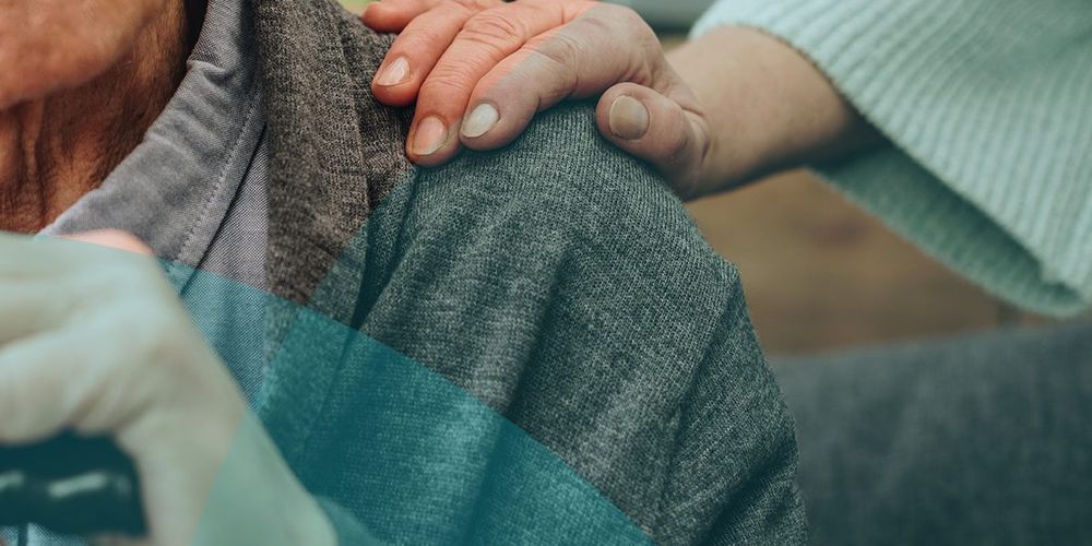 Elderly person in a grey sweater with a hand resting on their shoulder, showing comfort and support.