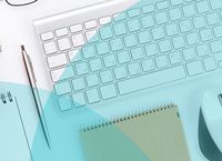 A desk setup with a keyboard, glasses, pen, notepad, and a mouse on a light blue surface.