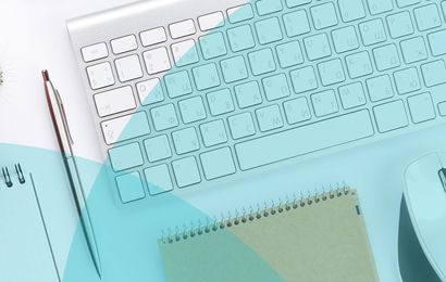 A desk setup with a keyboard, glasses, pen, notepad, and a mouse on a light blue surface.