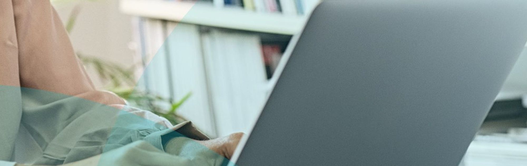 Person typing on a laptop at a desk, with books and plant in the background. Office setting.