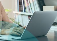 Person typing on a laptop at a desk, with books and plant in the background. Office setting.