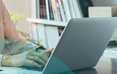 Person typing on a laptop at a desk, with books and plant in the background. Office setting.