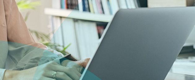 Person typing on a laptop at a desk, with books and plant in the background. Office setting.