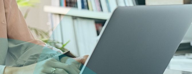 Person typing on a laptop at a desk, with books and plant in the background. Office setting.