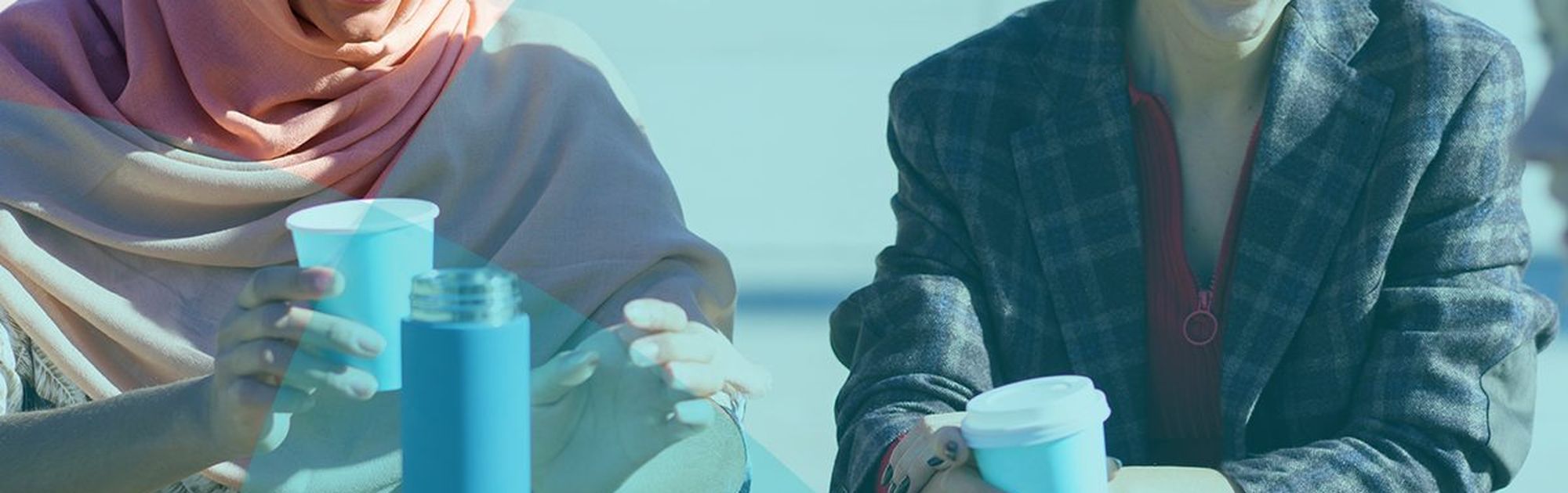 Two women sitting at an outdoor table, laughing and holding coffee cups, with a water bottle and smartphone nearby.
