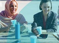 Two women sitting at an outdoor table, laughing and holding coffee cups, with a water bottle and smartphone nearby.