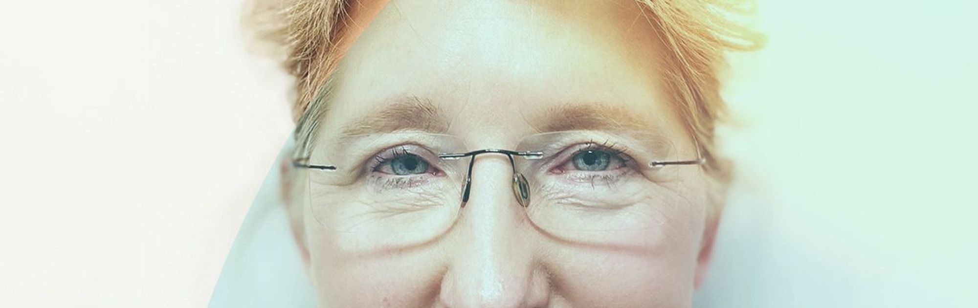 Smiling person with short blond hair and glasses, wearing a dark jacket. Soft, colorful overlay on the left side of the image.