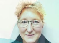 Smiling person with short blond hair and glasses, wearing a dark jacket. Soft, colorful overlay on the left side of the image.