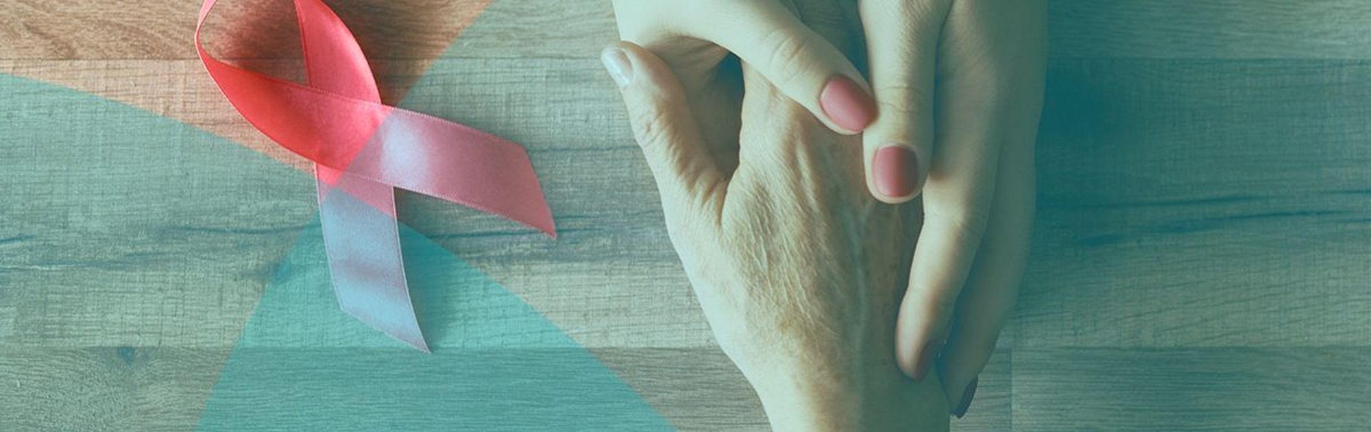 Two hands gently holding each other on a wooden surface next to a pink ribbon symbolizing breast cancer awareness.