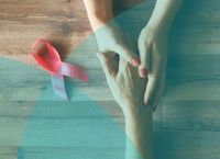 Two hands gently holding each other on a wooden surface next to a pink ribbon symbolizing breast cancer awareness.
