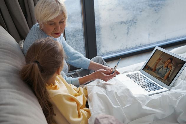 An elderly woman and a child talk with a nurse via a laptop in a small room. The elderly woman holds a thermometer.