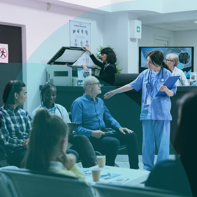 A nurse talks to a seated man in a hospital waiting room, surrounded by other patients and medical staff.