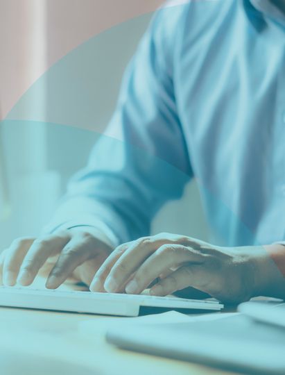 Person in a blue shirt typing on a keyboard at a desk, with soft lighting and a blurred background.