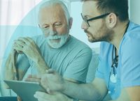 Elderly man with cane and younger doctor in scrubs discuss information on a tablet in a well-lit room.