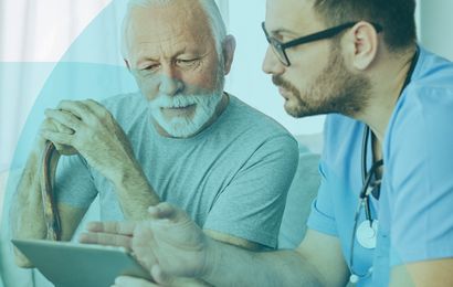 Elderly man with cane and younger doctor in scrubs discuss information on a tablet in a well-lit room.