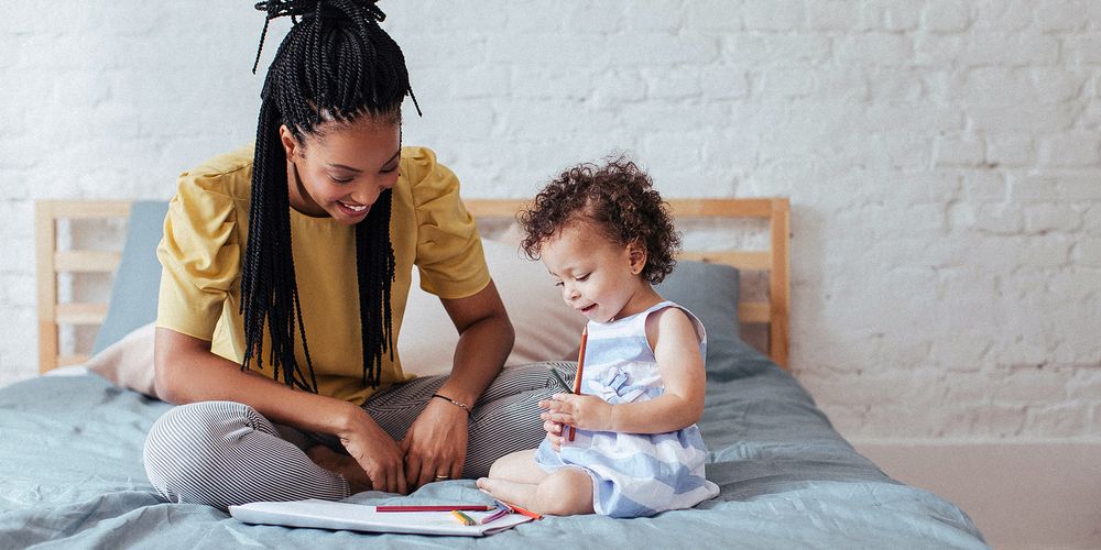 A woman and a young child sit on a bed drawing together, surrounded by a cozy bedroom setting with a white brick wall.