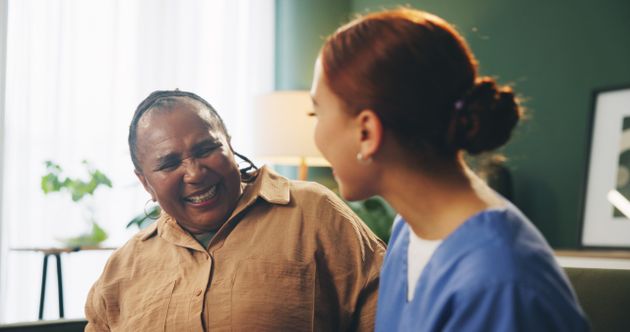 Elderly person smiling with a caregiver in a blue uniform in a cosy room with plants and soft lighting.