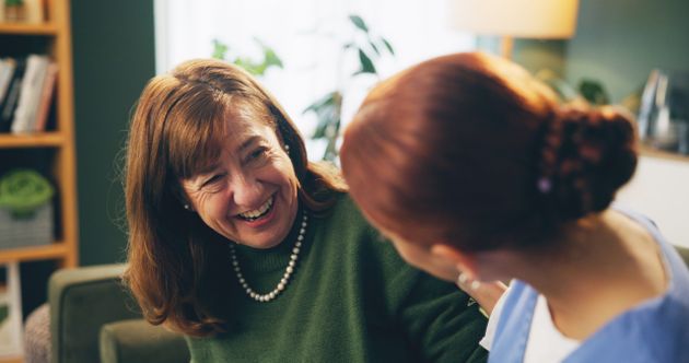Two women smiling and engaging in a lively conversation in a cozy room, with bookshelves and plants in the background.