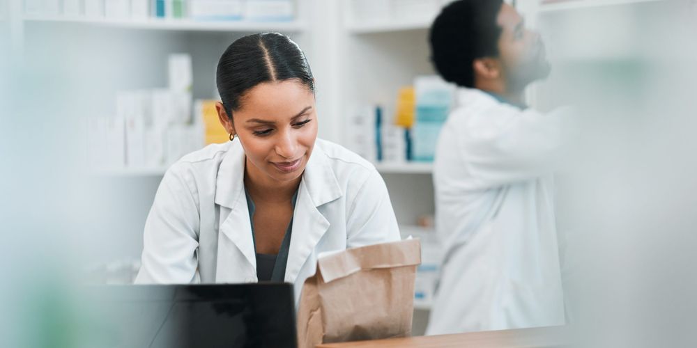 Two pharmacists in lab coats work in a pharmacy. One checks inventory on a laptop, while the other reaches for a medication on the shelves.