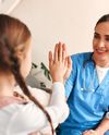 A smiling nurse in blue scrubs high-fives a young girl, with a seated woman nearby, in a warmly lit room with plants.