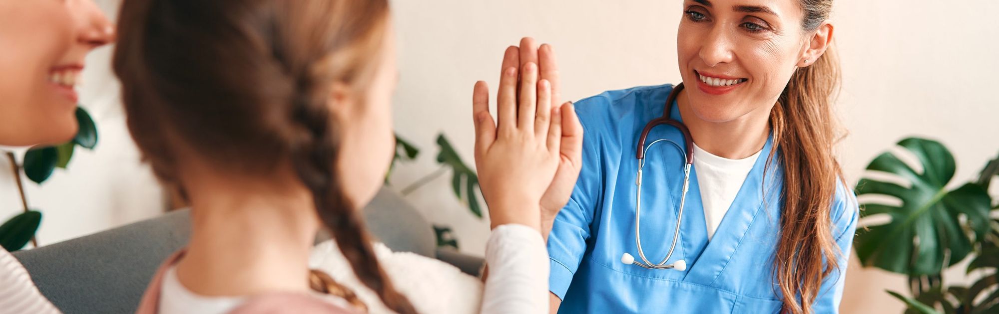 A smiling nurse in blue scrubs high-fives a young girl, with a seated woman nearby, in a warmly lit room with plants.