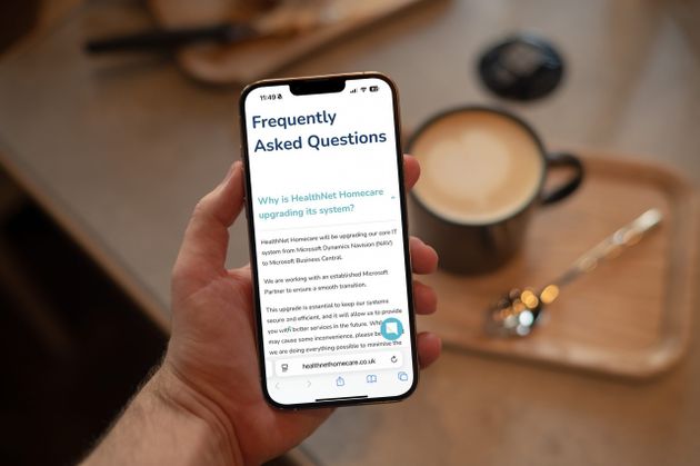 A person holding a smartphone displaying a FAQ webpage, with a coffee cup and saucer in the background.