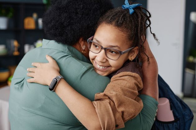 A smiling girl with glasses and a blue bow in her braided hair hugs someone wearing a green shirt, expressing warmth and affection.