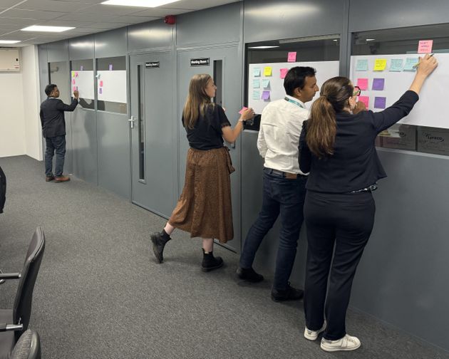 Four people write on sticky notes attached to paper on office doors, engaging in a collaborative brainstorming session.
