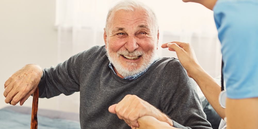 Close-up of an older adult seated indoors holding a walking stick while a healthcare professional gently supports their arm during a home visit.