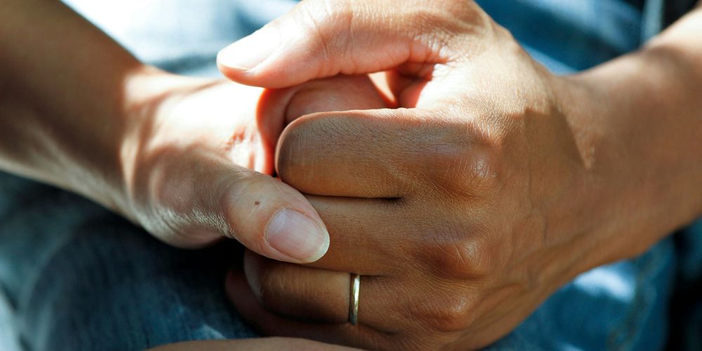 Two hands gently hold each other, with one wearing a ring, symbolizing support and connection.