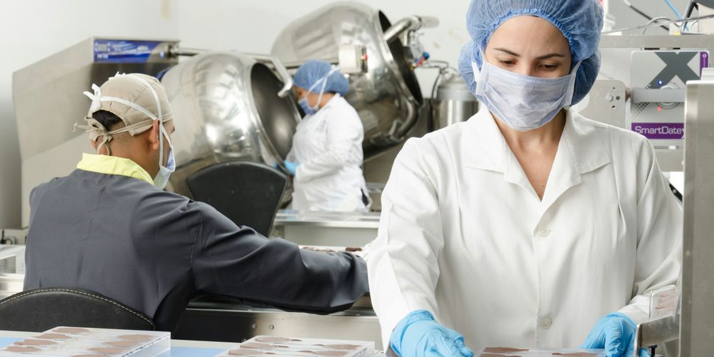 Workers in protective gear handling chocolate molds in a factory setting, with machinery in the background.