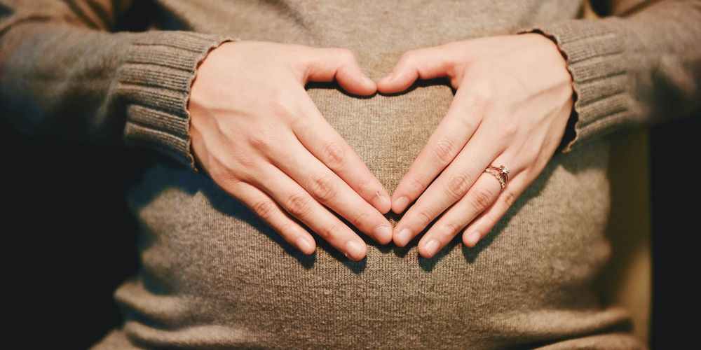 Pregnant person wearing a gray sweater holds hands in a heart shape over their belly.