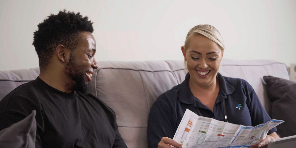 A man and a woman sit on a couch, smiling while looking at a brochure.